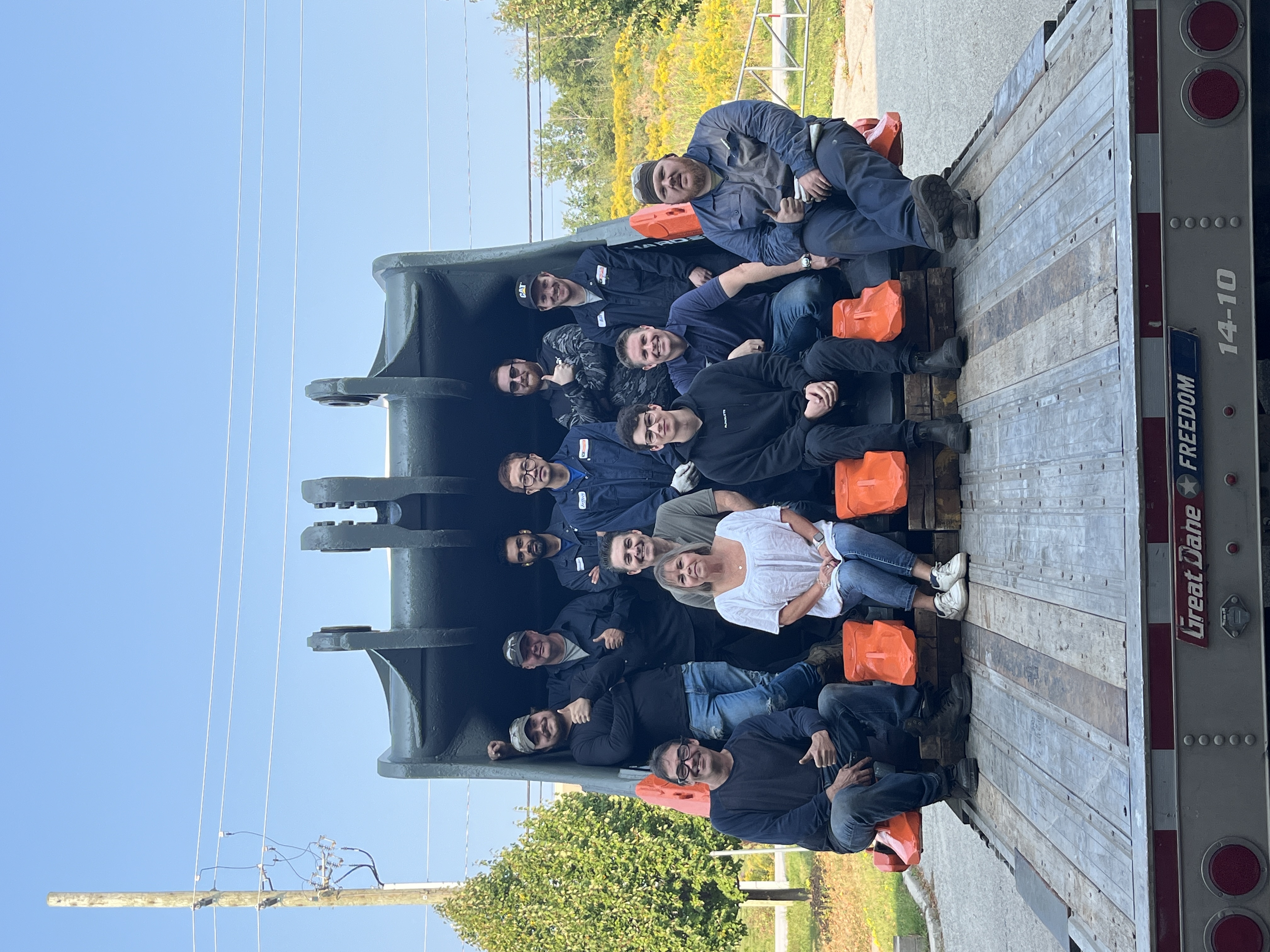 Grupo de trabajadores posando dentro de un gran balde de excavadora cargado en un remolque de plataforma, con dientes naranjas sobre la cubierta frente a ellos