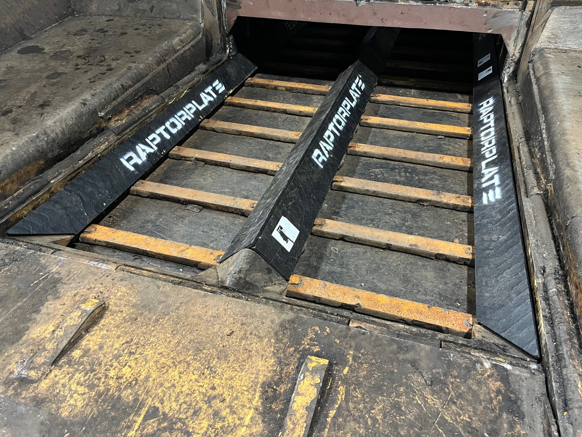 Tres barras de desgaste RaptorPlate negras instaladas sobre una cuadr&iacute;cula de barras desgastadas con bordes naranjas en el piso de un chute