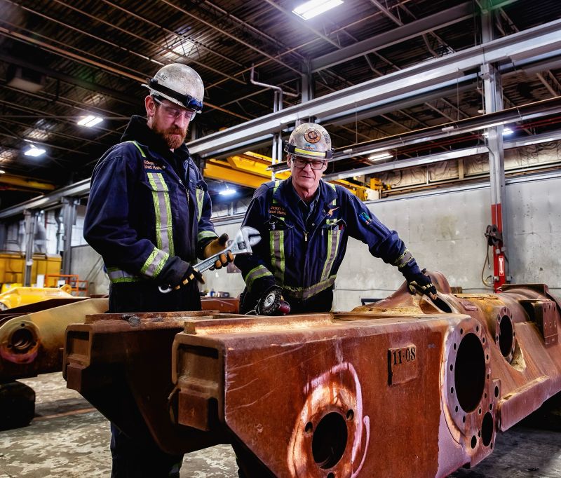 Dos trabajadores con overoles azul marino y cascos inspeccionando una gran fundici&oacute;n de acero oxidada en un taller, uno de ellos con una llave