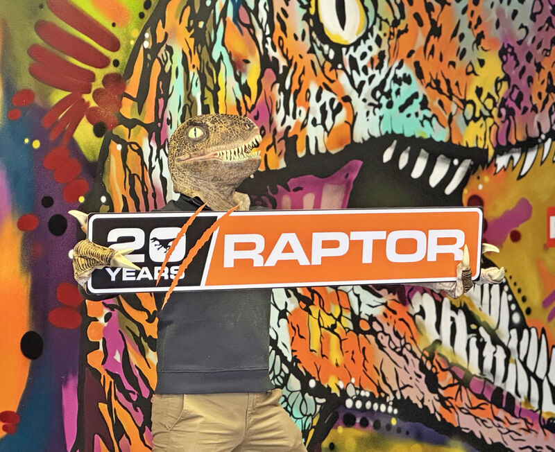 Persona con m&aacute;scara de dinosaurio sosteniendo un letrero de Raptor 20 Years frente a un mural colorido de un raptor