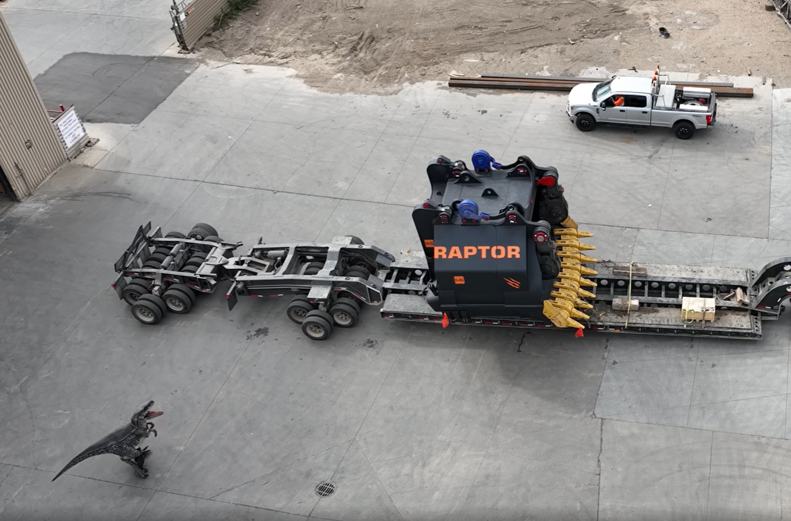 Vista a&eacute;rea de un gran balde RAPTOR negro con dientes amarillos que se transporta en un remolque, con un disfraz de dinosaurio sobre el pavimento y una camioneta Raptor cerca